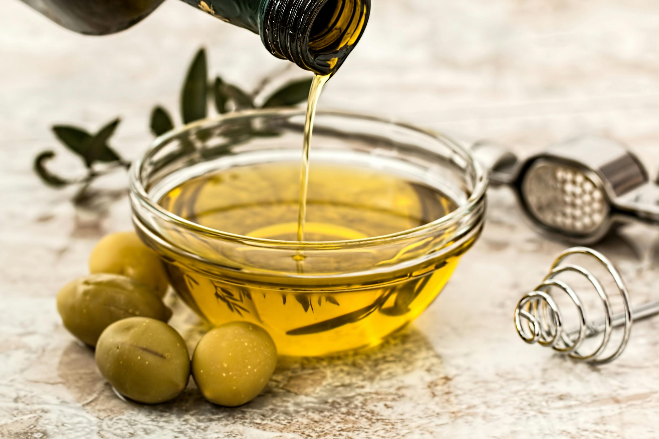 Close-up of olive oil being poured into a glass bowl surrounded by fresh olives and kitchen tools.